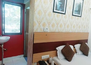 Habitación de hotel con cabecera de madera, cama con almohadas blancas y marrones, una mesita de noche pequeña y ventana con vistas a árboles.