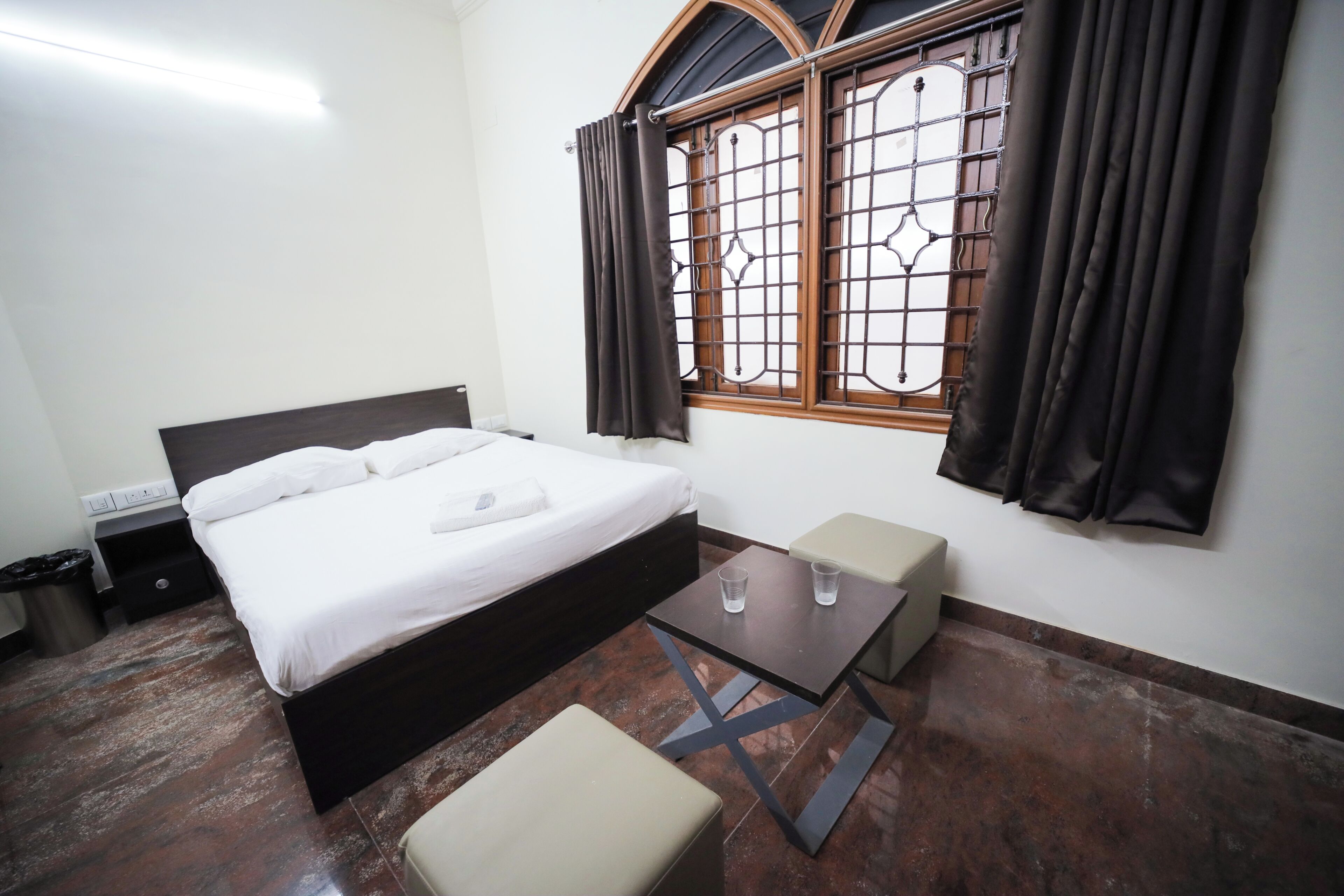 Una habitación de hotel con cama, una mesita, un banco, una ventana con cortinas y un cesto de basura.