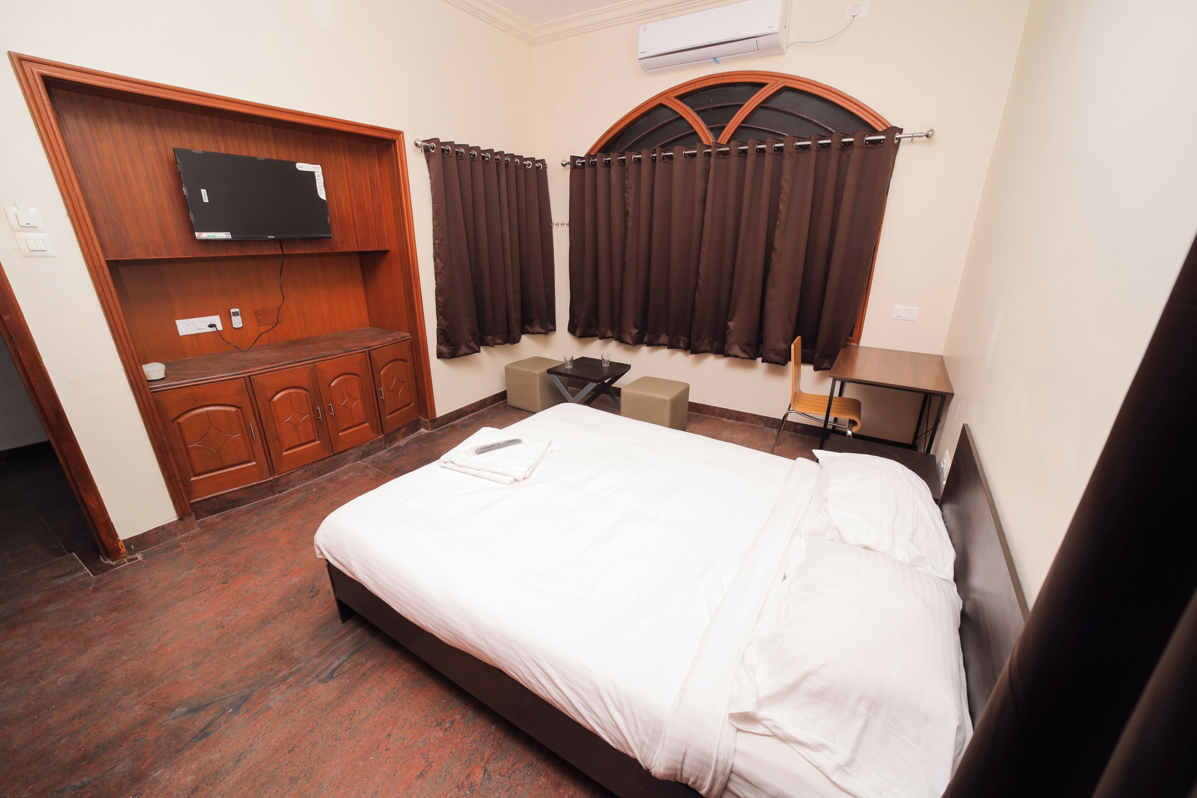 A hotel room with a bed, a TV mounted on a wooden cabinet, a desk with a chair, and a window with curtains.