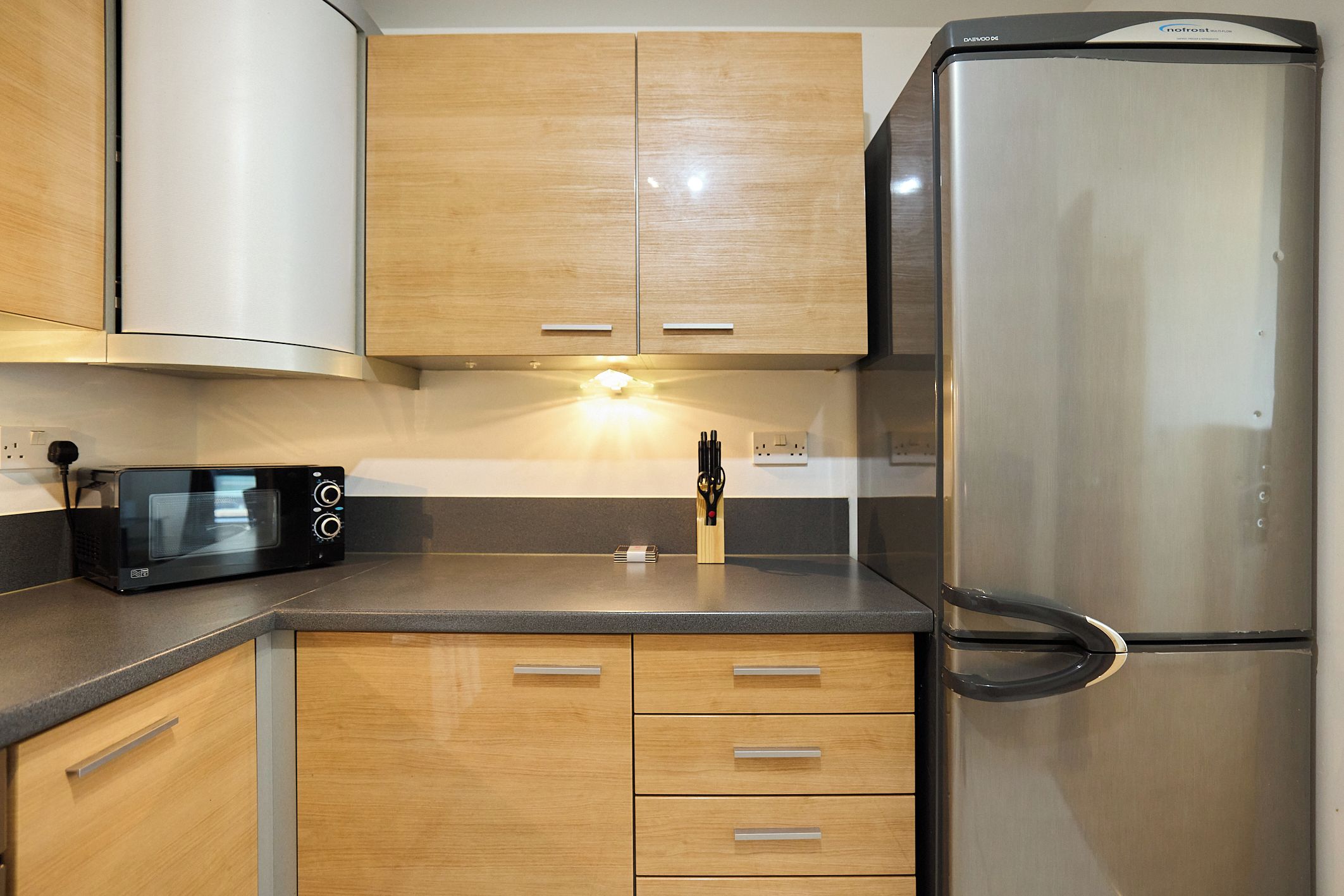 Una cocina con armarios de madera, un refrigerador de acero inoxidable, un microondas y un bloque de cuchillos.