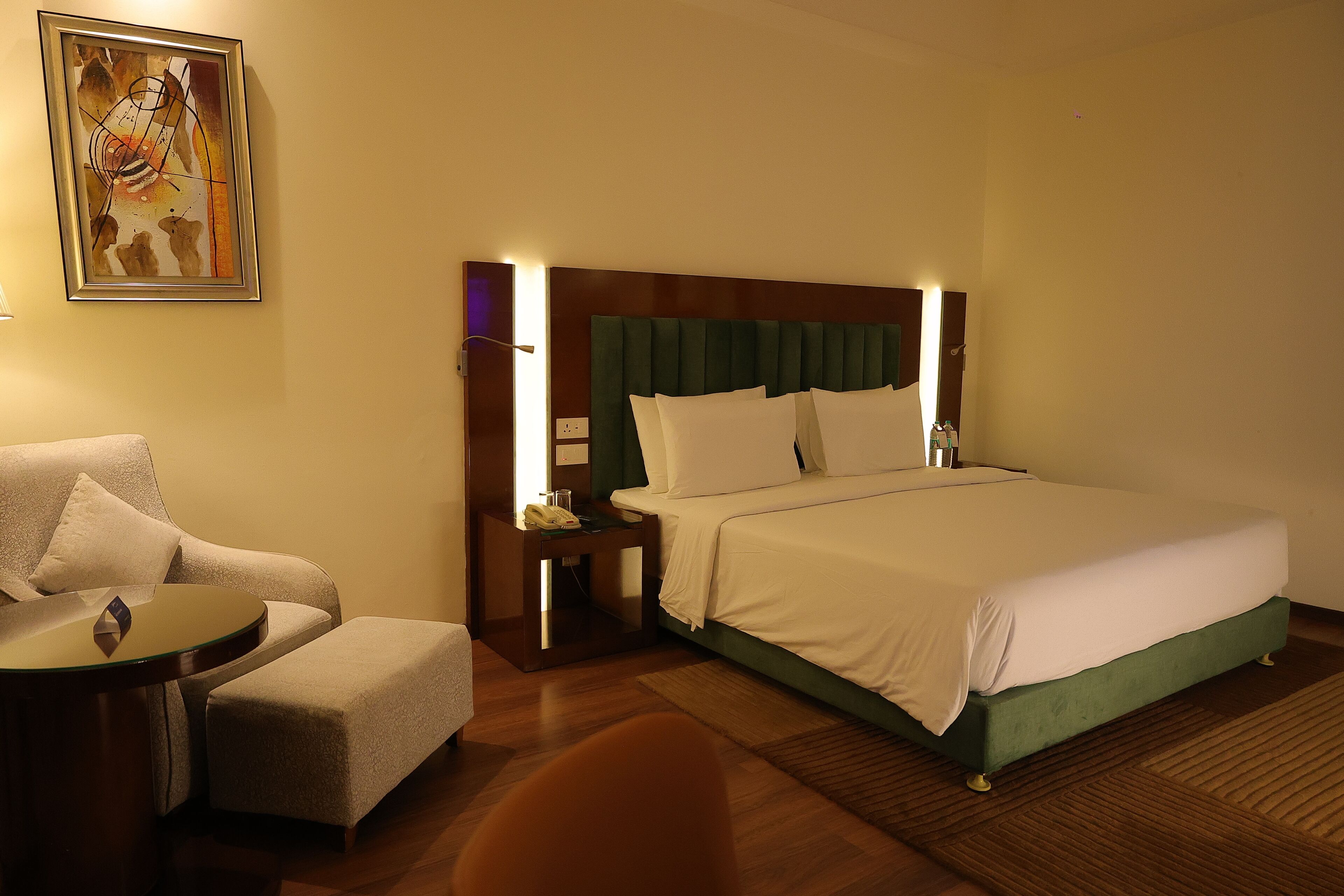 A hotel room with a bed, a chair, a small table, and a painting on the wall.