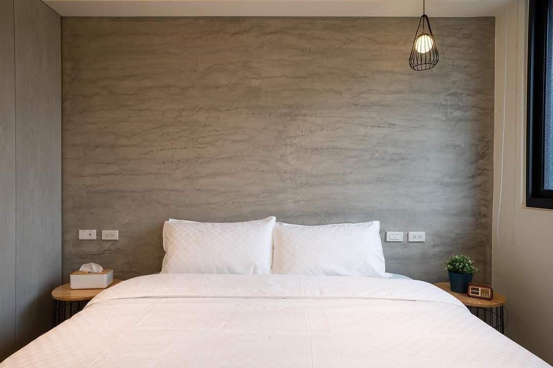 A bedroom with a large bed, a wall-mounted light fixture, bedside tables with a radio and a plant, and a window on the right.