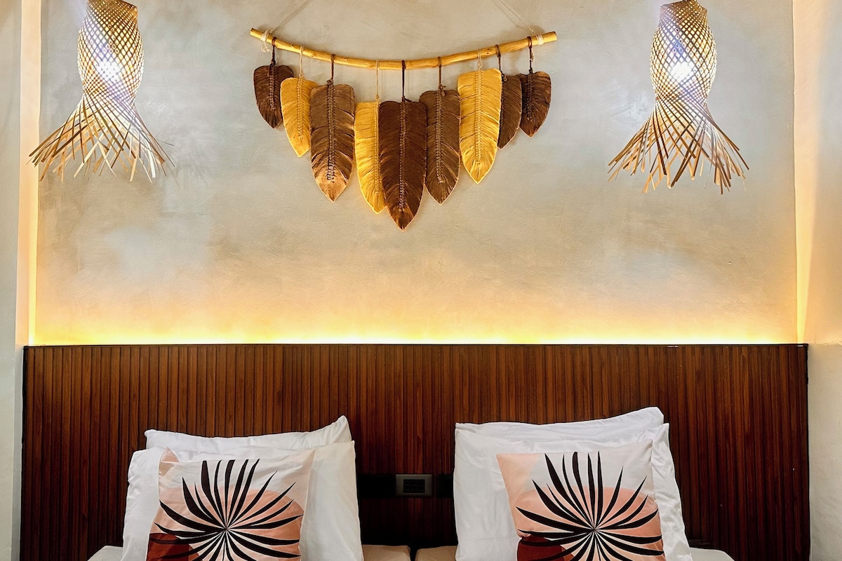 Habitación de hotel con una cama, cabecera de madera, dos almohadas con diseño de hojas y una lámpara de pared con motivos vegetales.