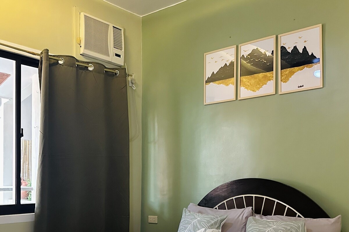 A neatly made bed with pillows and rolled towels, a window with a curtain, and wall-mounted air conditioning.
