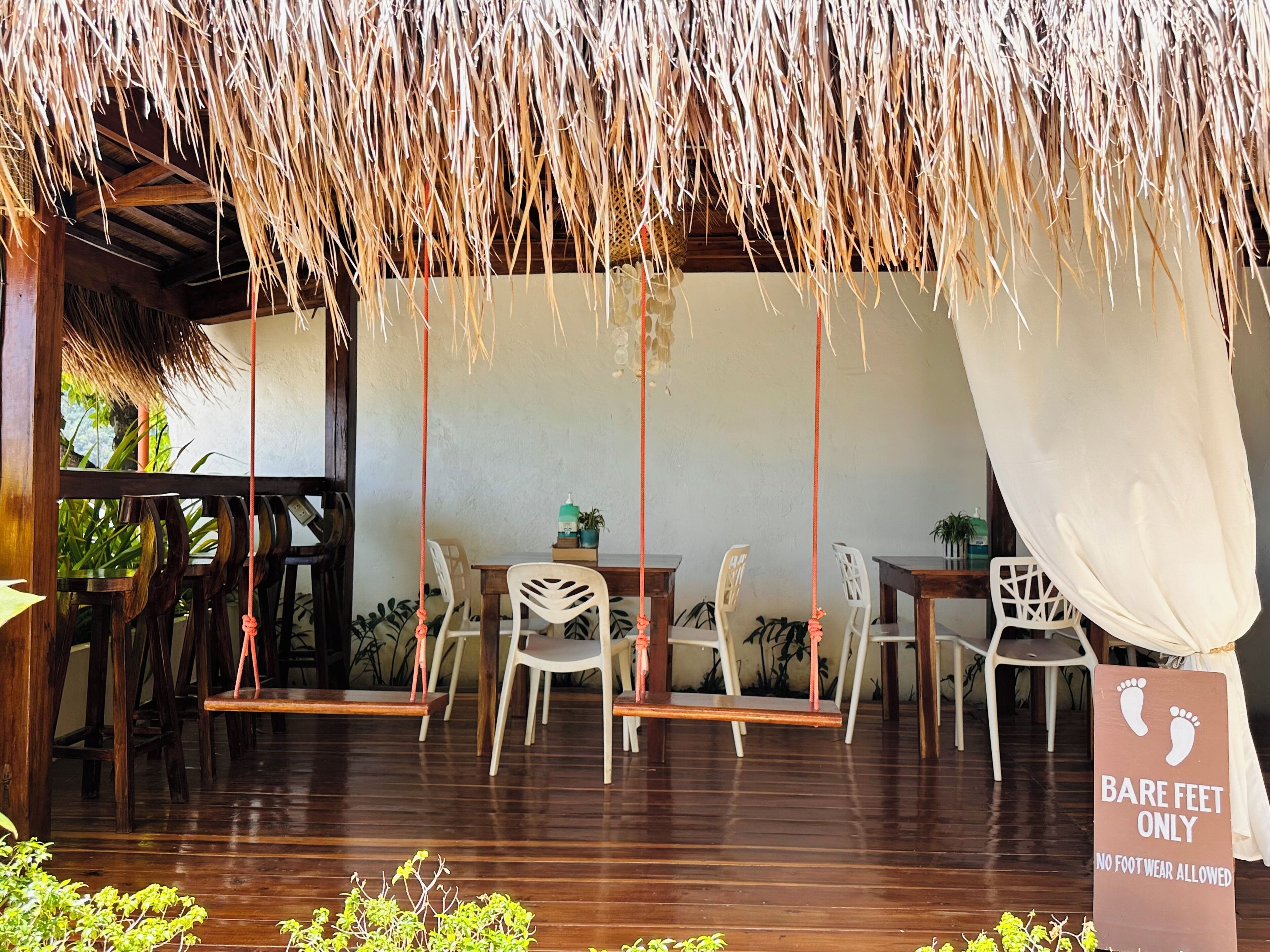 Un espace extérieur aménagé avec des tables et des chaises en bois, un toit de chaume et un panneau indiquant : «Pieds nus uniquement».