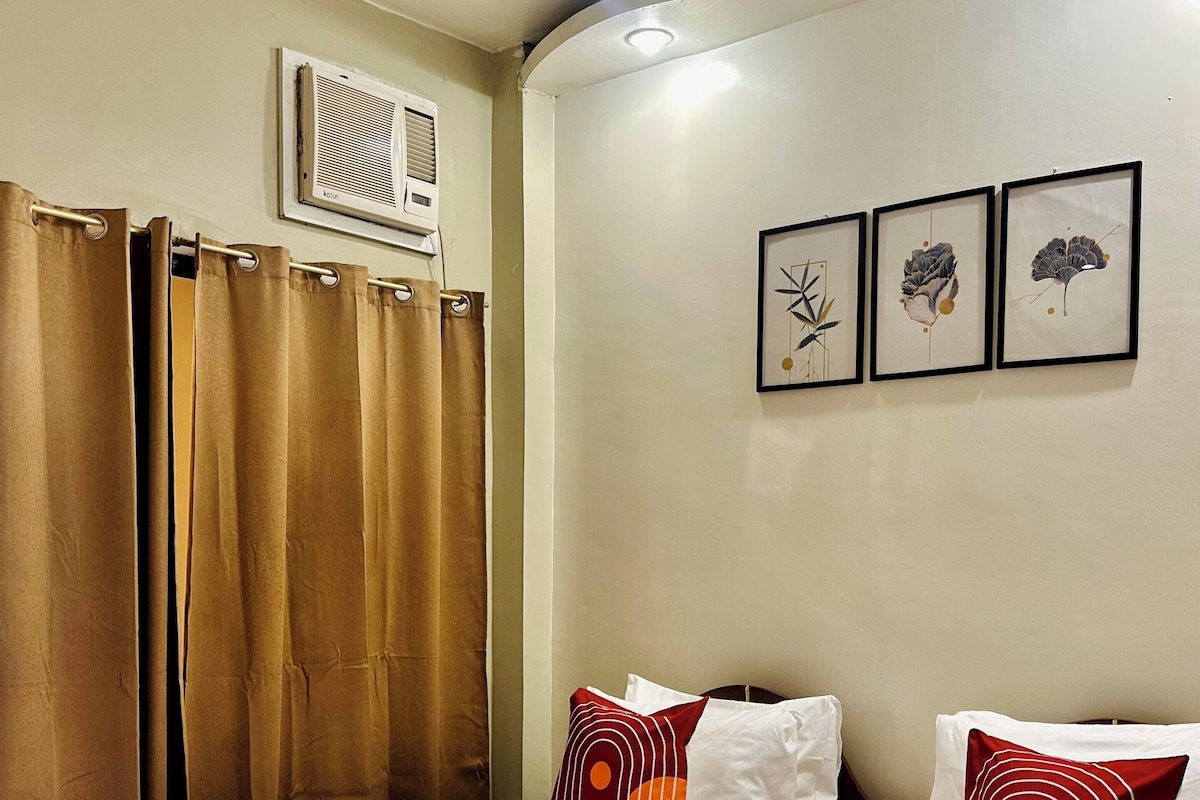A hotel room with two beds, brown curtains, a ceiling fan, an air conditioning unit, and three framed wall art pieces.
