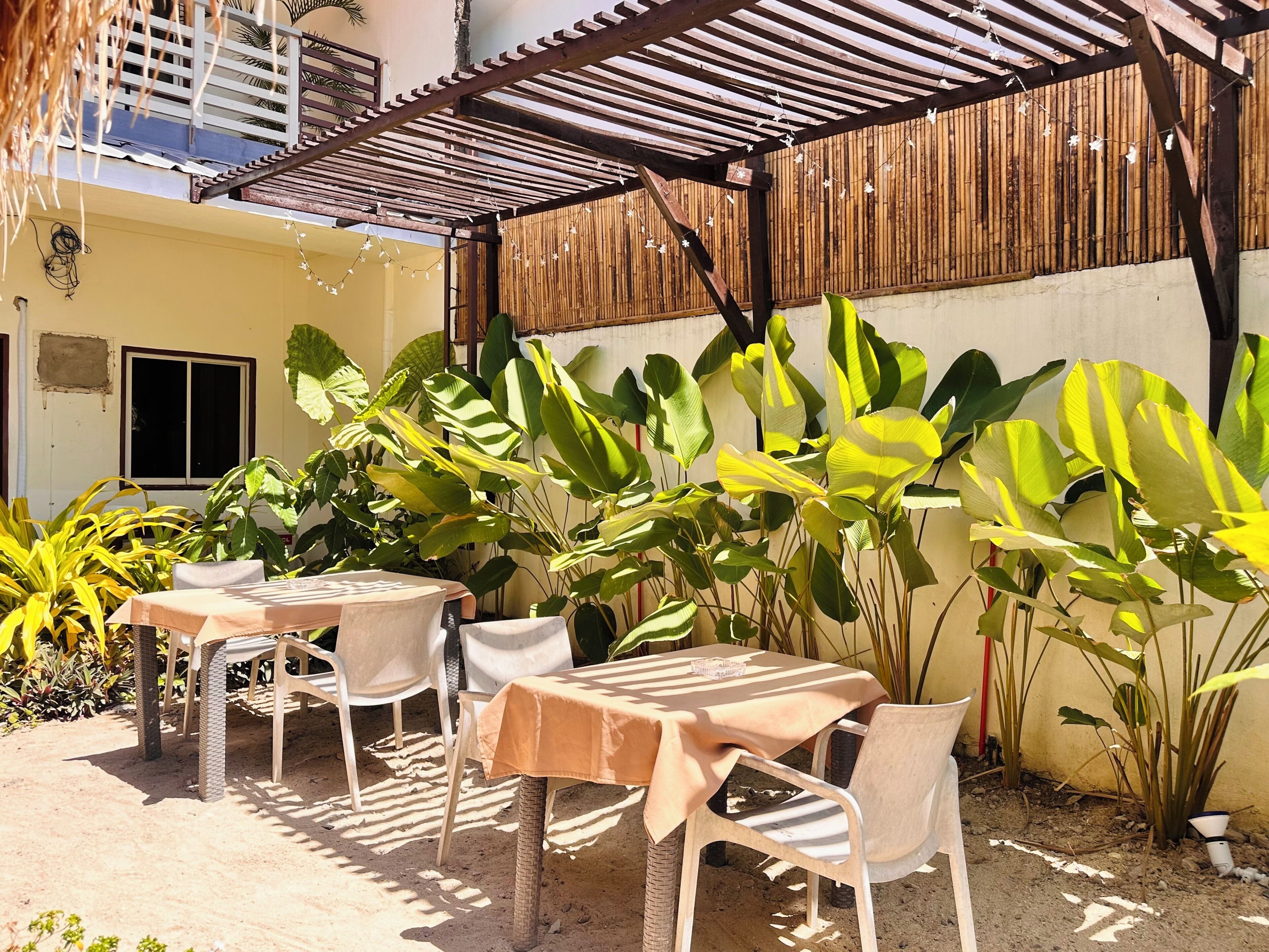 An outdoor patio with tables and chairs, surrounded by large green plants and a wooden fence.