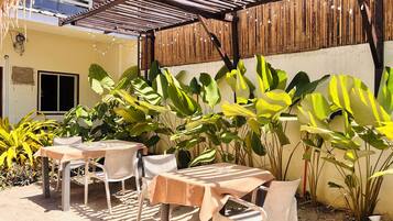 An outdoor patio with tables and chairs, surrounded by large green plants and a wooden fence.