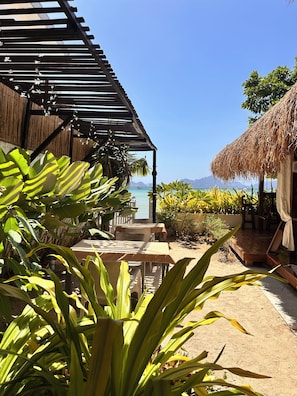 A beachside patio with tables and chairs, a thatched-roof structure, and a view of the ocean. - Panari Lodge El Nido  (El Nido)