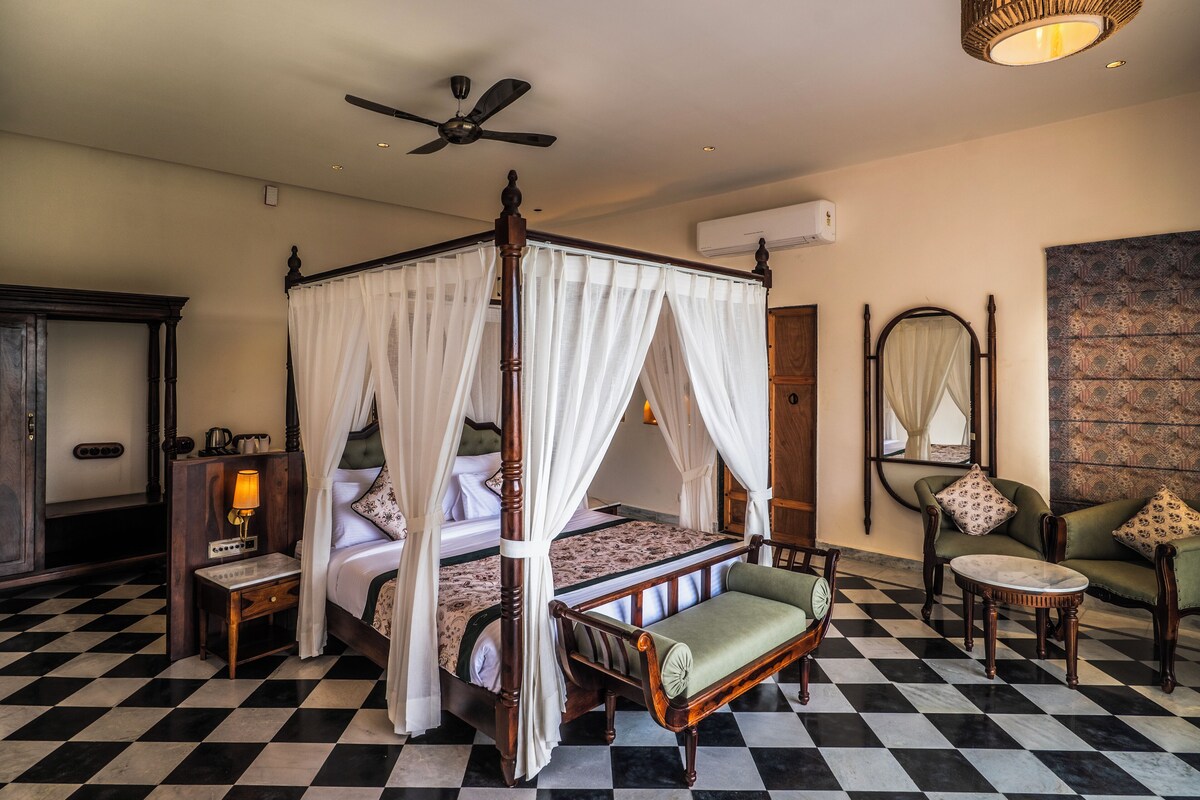 A four-poster bed with a canopy, a bedside table with a lamp, a chair, and a small round table.