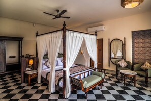 A four-poster bed with a canopy, a bedside table with a lamp, a chair, and a small round table. - The Talhati (Kumbhalgarh)
