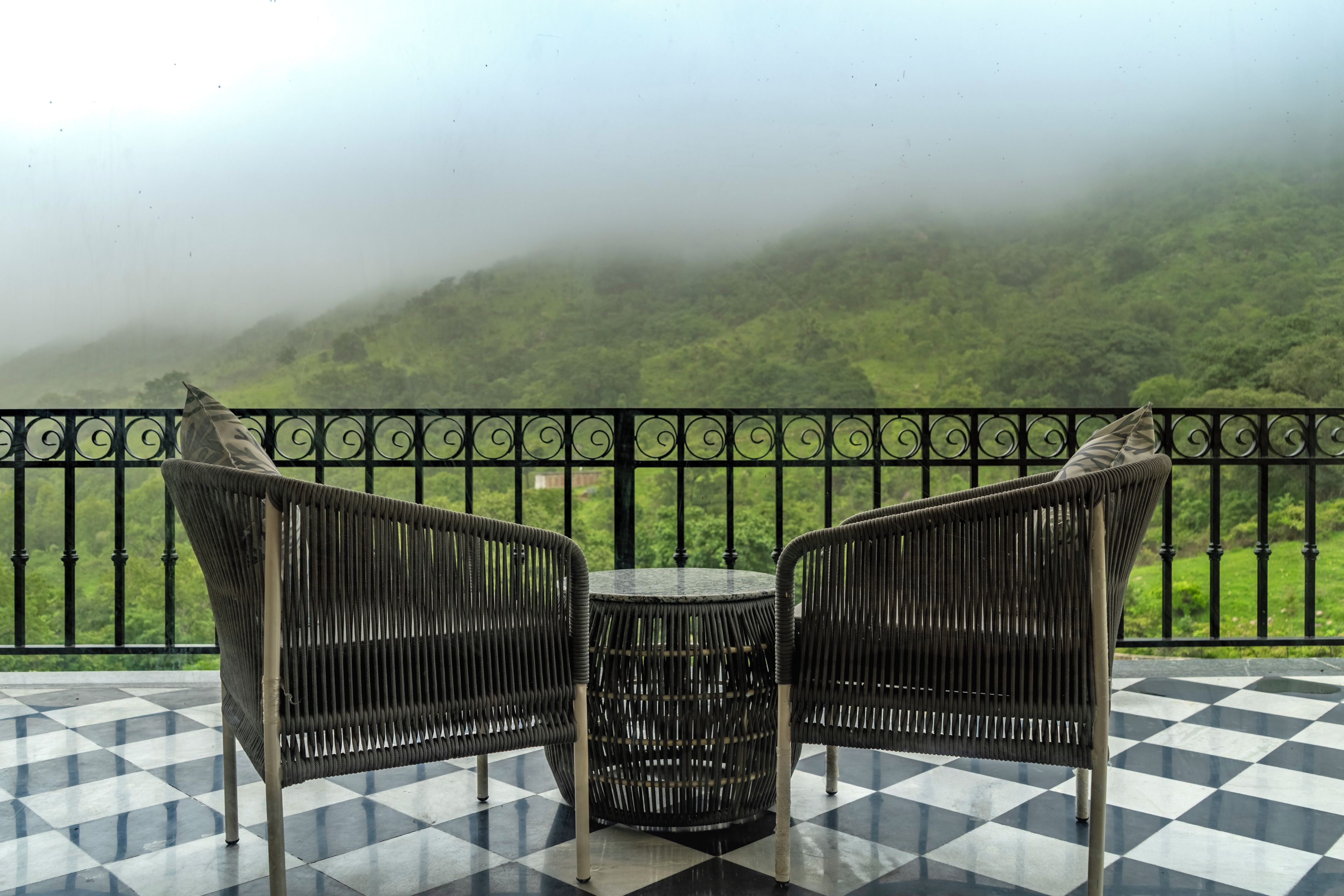 A balcony with two wicker chairs and a small round table, overlooking a lush green landscape with hills in the background.
