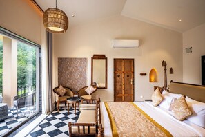 Habitación de hotel con piso a cuadros, cama con colcha estampada, zona de estar con sillas y mesa, ventana grande con vista a la vegetación y unidad de aire acondicionado en la pared.