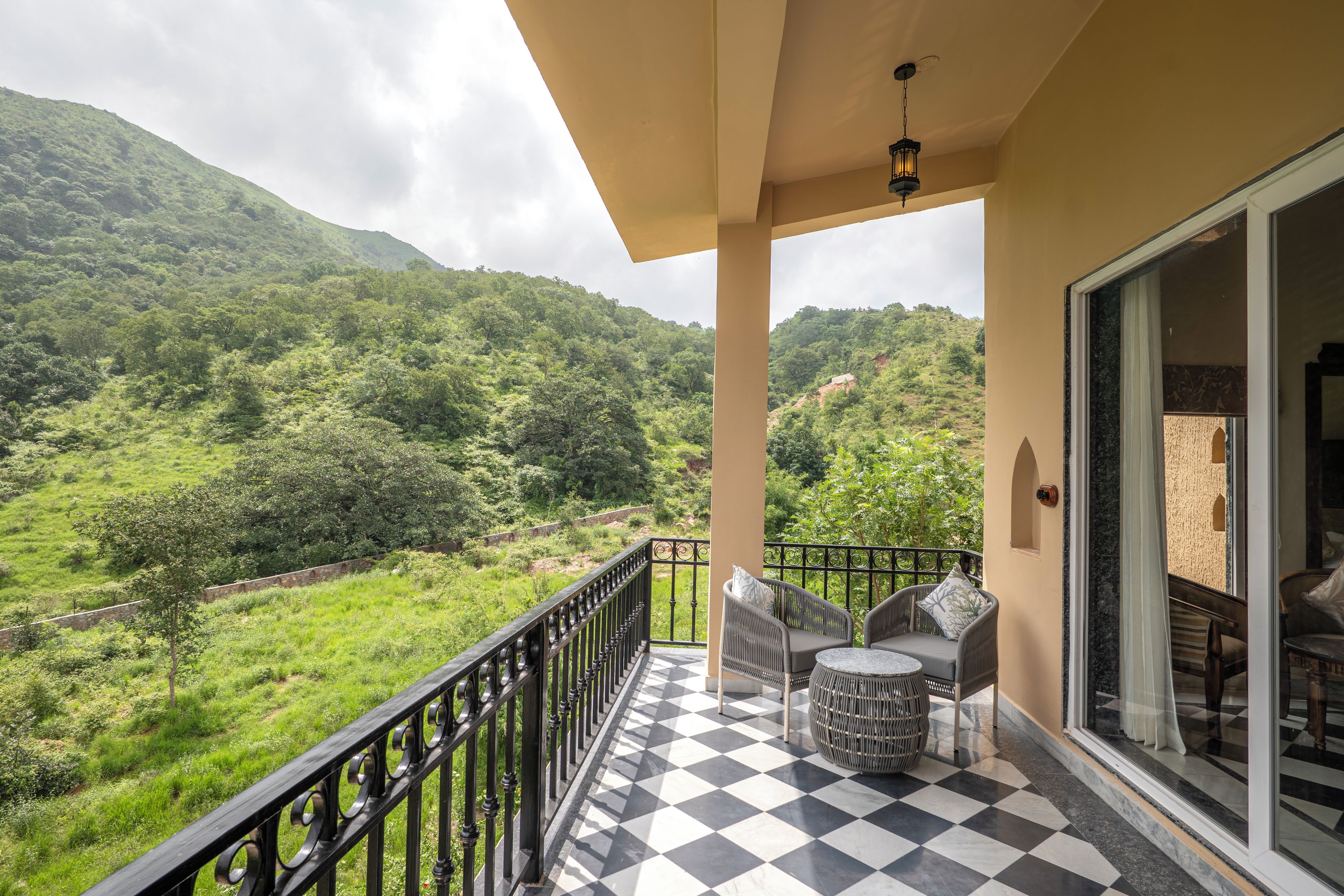A balcony with a checkered floor, two wicker chairs, a small round table, and a view of a lush green valley with hills.
