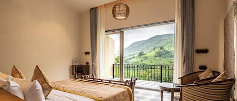 Quarto de hotel com cama, uma cadeira, uma mesa e vista para uma paisagem montanhosa através de uma grande janela.