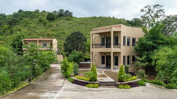 A two-story building with a balcony, surrounded by lush greenery and a hillside.
