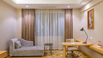 A hotel room with a bed, a sofa, a desk, a chair, a small table, and a painting on the wall.