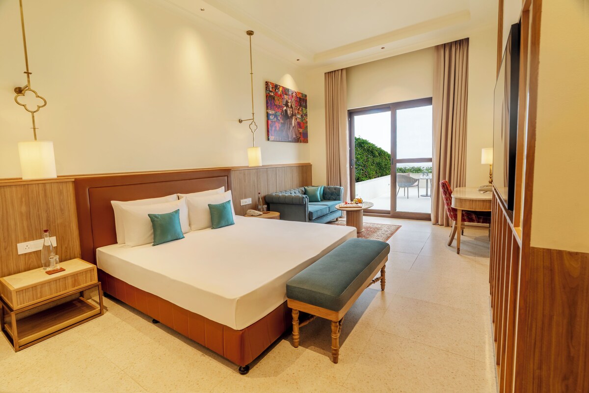 A hotel room with a large bed, bedside tables, a bench, a small table with a fruit bowl, a sofa, and a view of the outdoors.