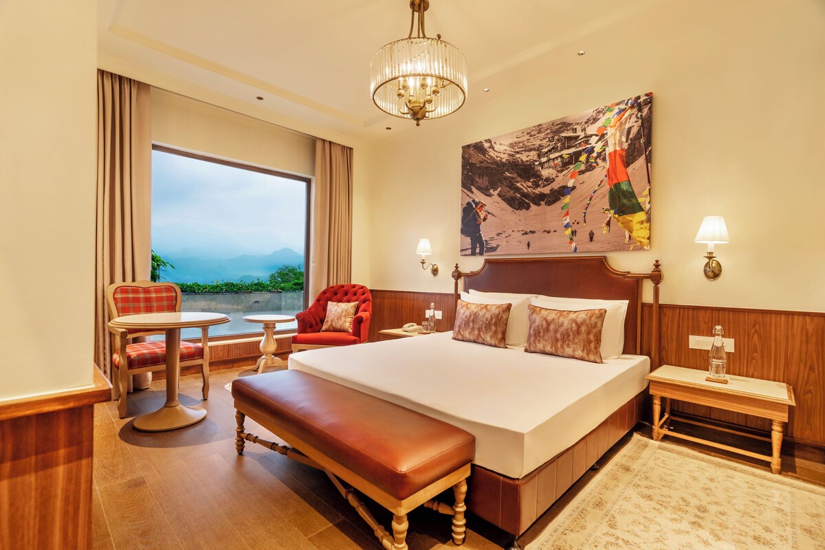 A hotel room with a large bed, a bench, a chair, a table, a lamp, and a painting of a mountainous landscape.
