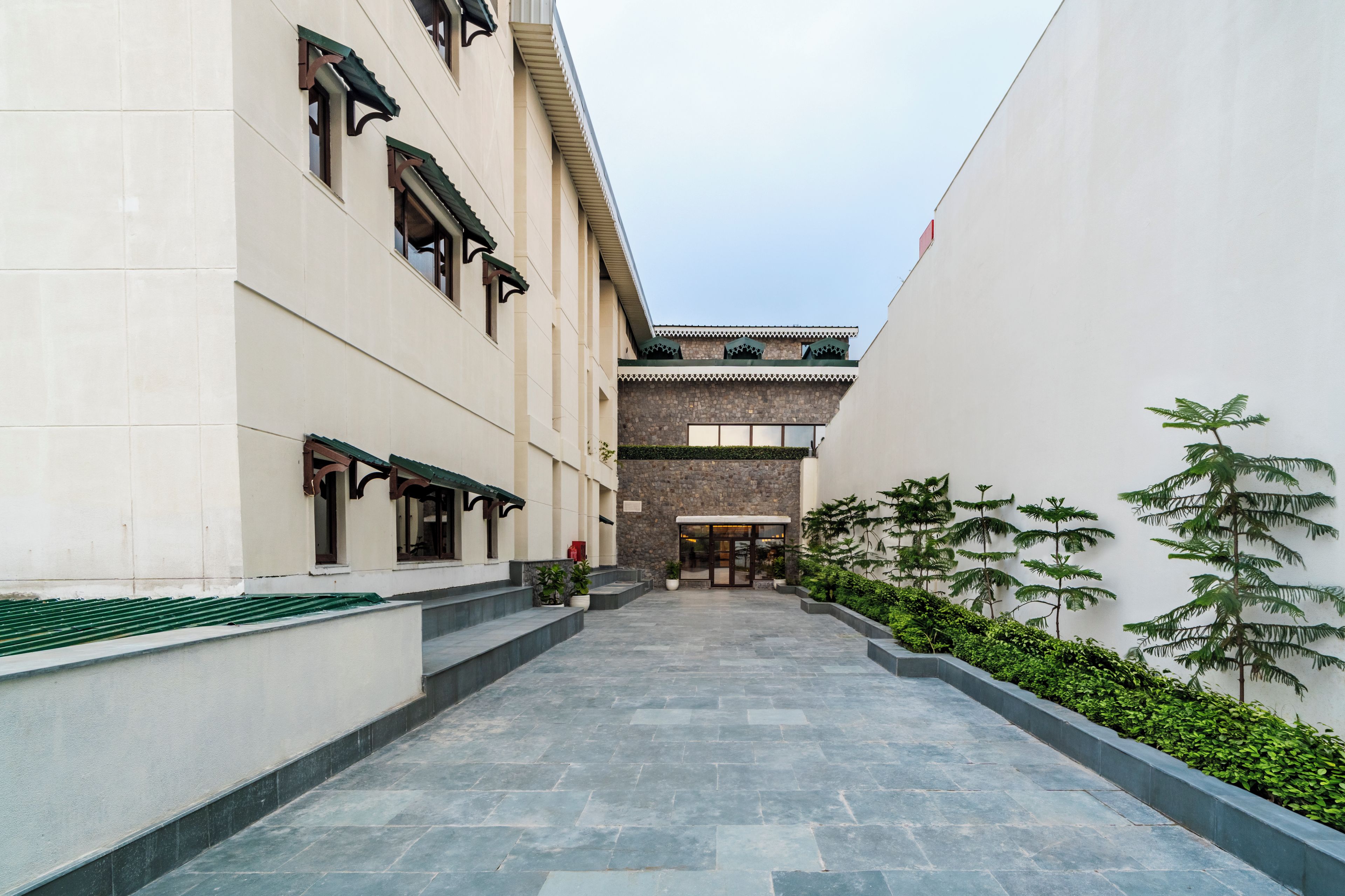 A corridor with a stone floor, a building with a traditional facade, and a small garden area with potted plants.