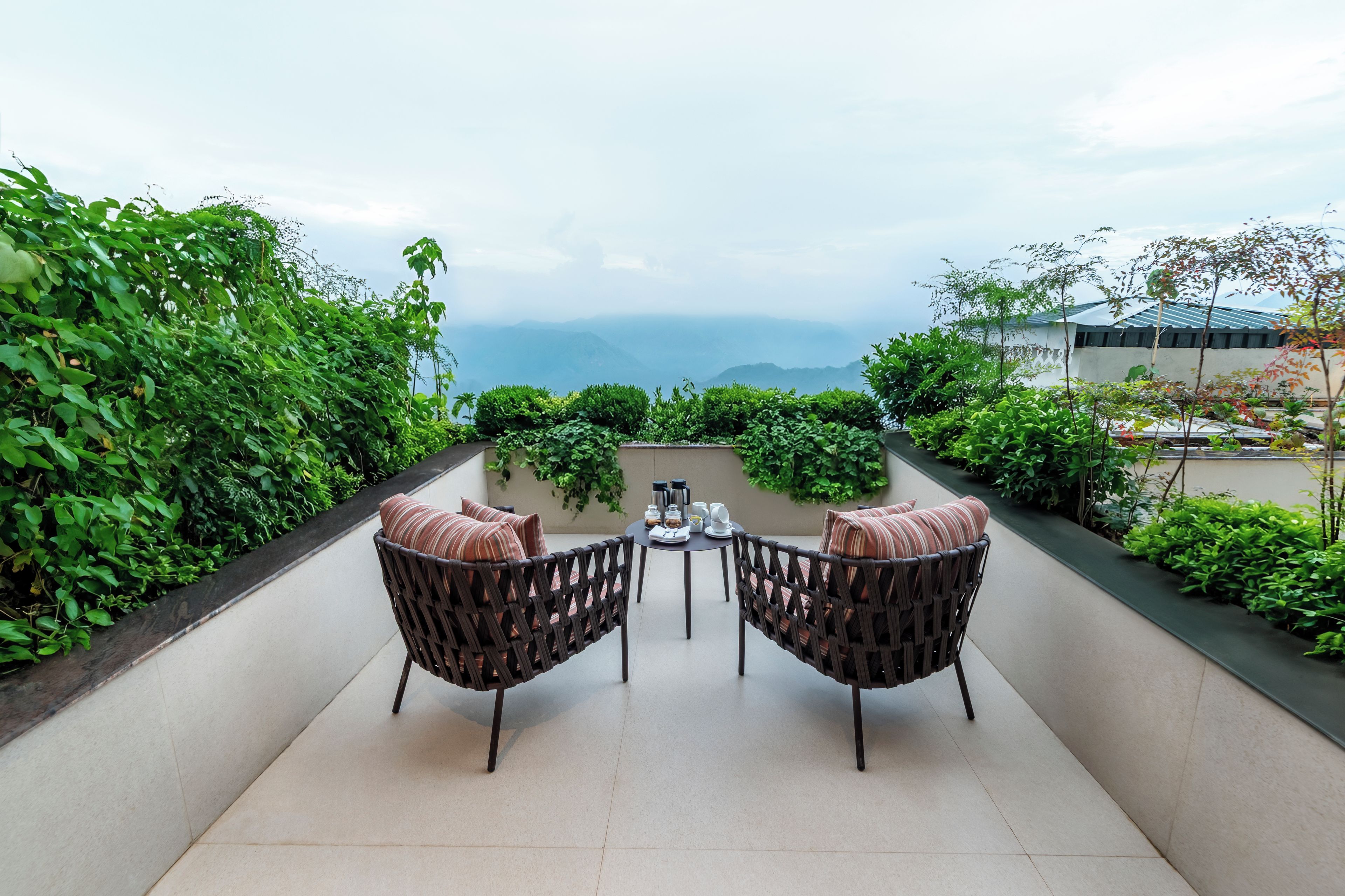 Eine Terrasse mit zwei Korbstühlen und einem Tisch, umgeben von üppiger Vegetation mit Blick auf eine gebirgige Landschaft.