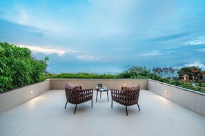 Una terraza con dos sillas de mimbre y una mesita pequeña, con vistas a un jardín exuberante y montañas lejanas.