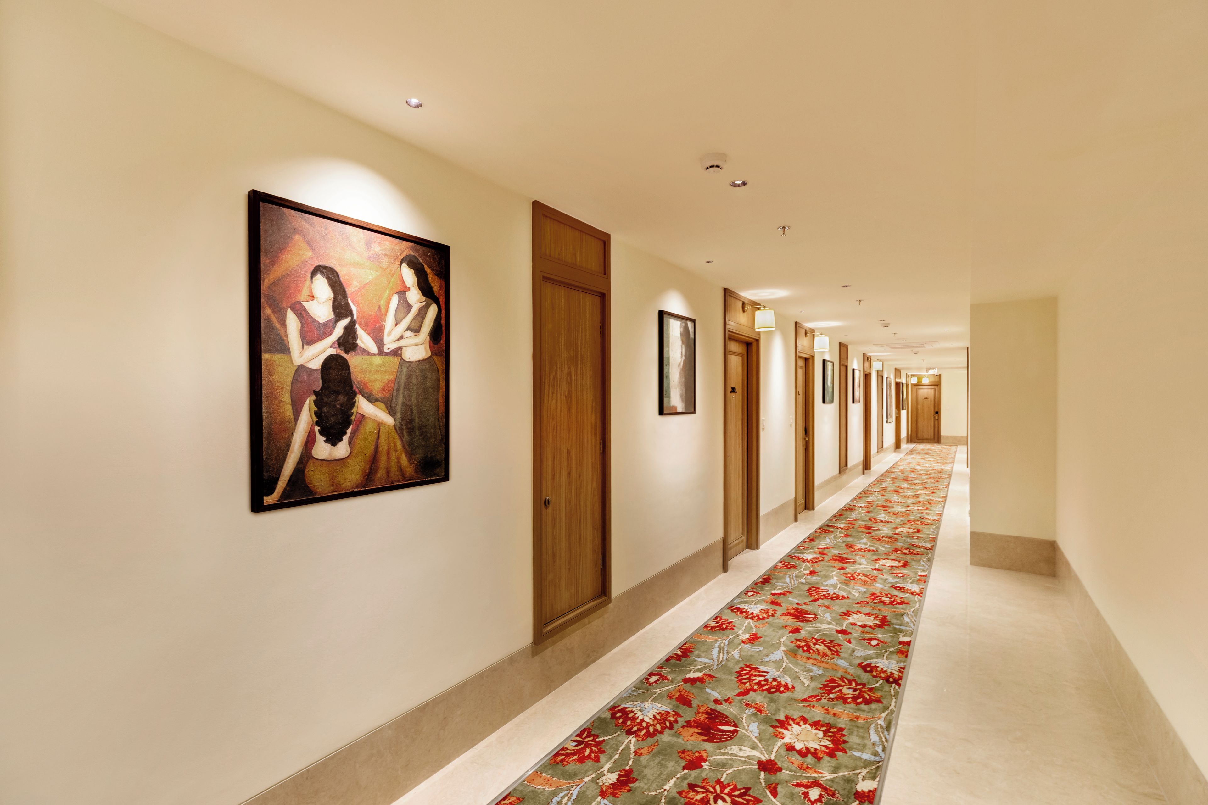 A hotel hallway with a floral carpet, wooden doors, and framed artwork on the walls.