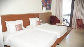 Quarto de hotel com duas camas, uma poltrona vermelha, uma mesinha com uma garrafa e um copo, e uma varanda com uma cadeira.