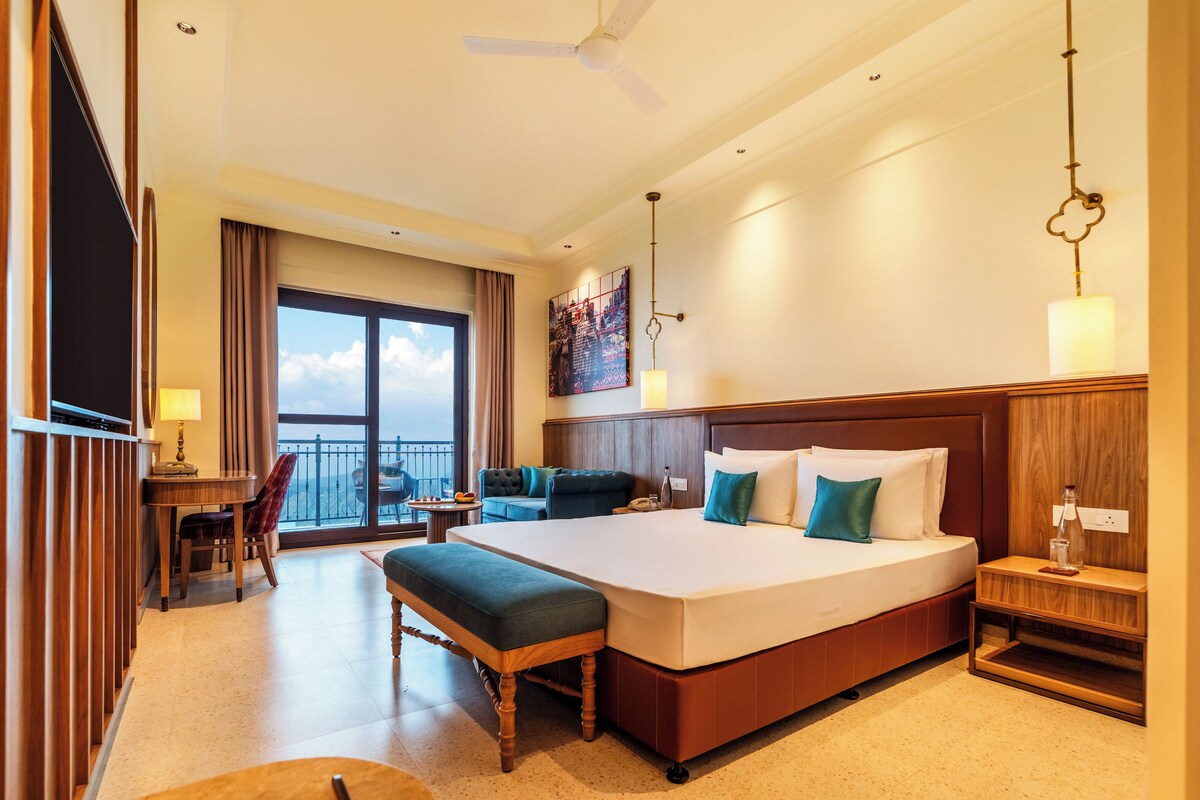 A hotel room with a large bed, a desk with a chair, a balcony with a view, and a ceiling fan.