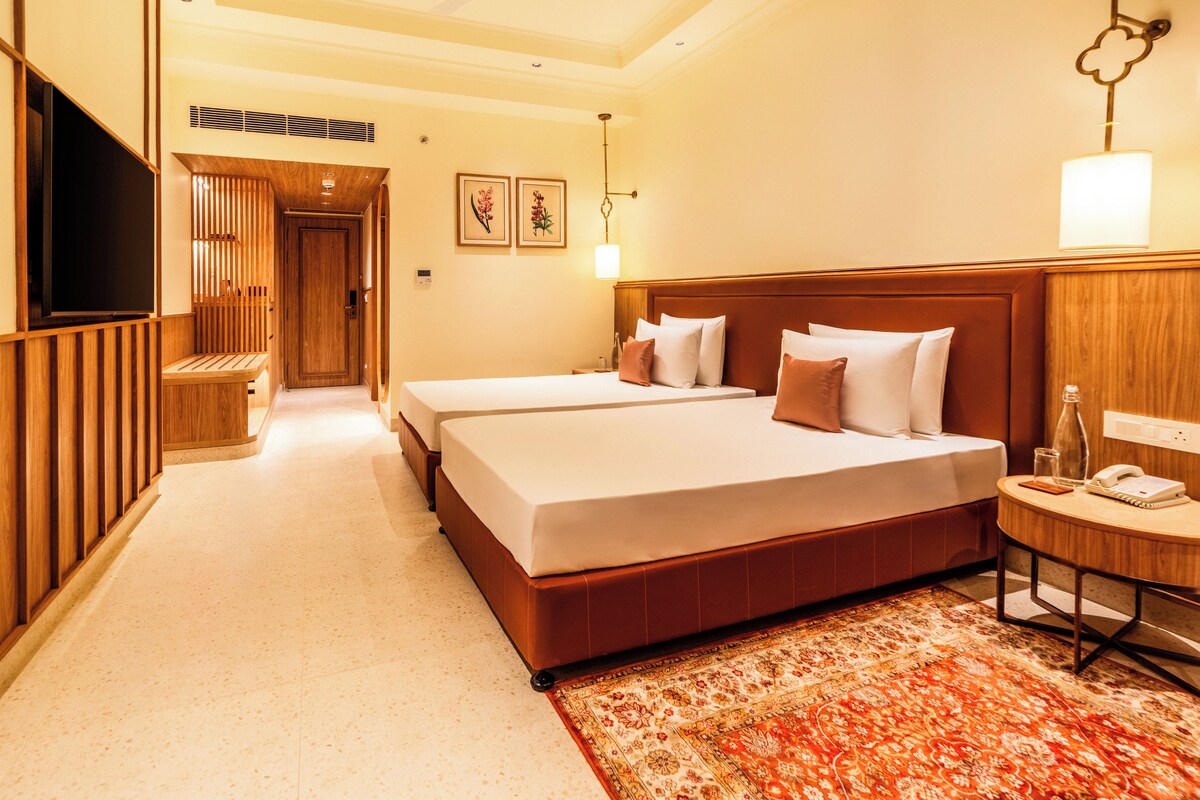 A hotel room with two beds, a television, a bedside table with a lamp, and a carpeted floor.
