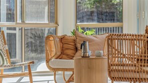 A wicker sofa with cushions, a metal coffee pot, and a cup on a small table, with a window in the background.