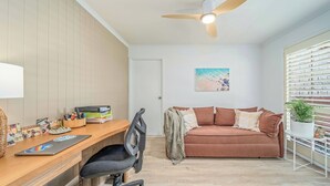 Una habitación con un escritorio de madera, una silla de oficina negra, un sofá marrón, un ventilador de techo y una ventana con persianas.