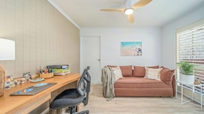A room with a wooden desk, a black office chair, a brown sofa, a ceiling fan, and a window with blinds.