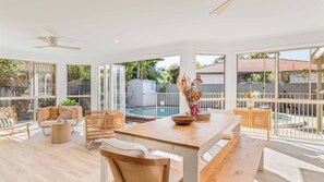 A spacious indoor area with a wooden pool table, wicker furniture, and large windows offering views of a backyard with a pool and greenery.