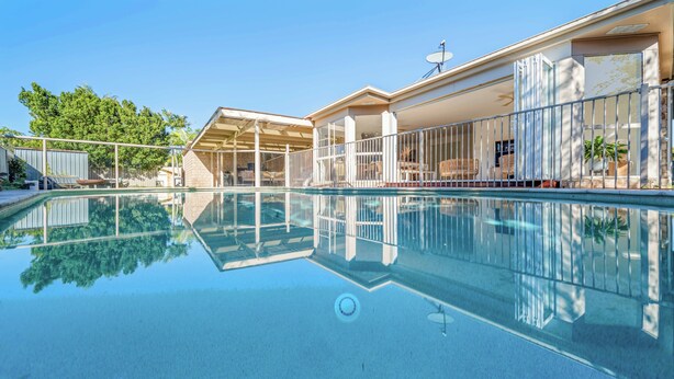 Piscina transparente com fundo de vidro, rodeada por um terraço coberto e mobiliário de exterior.