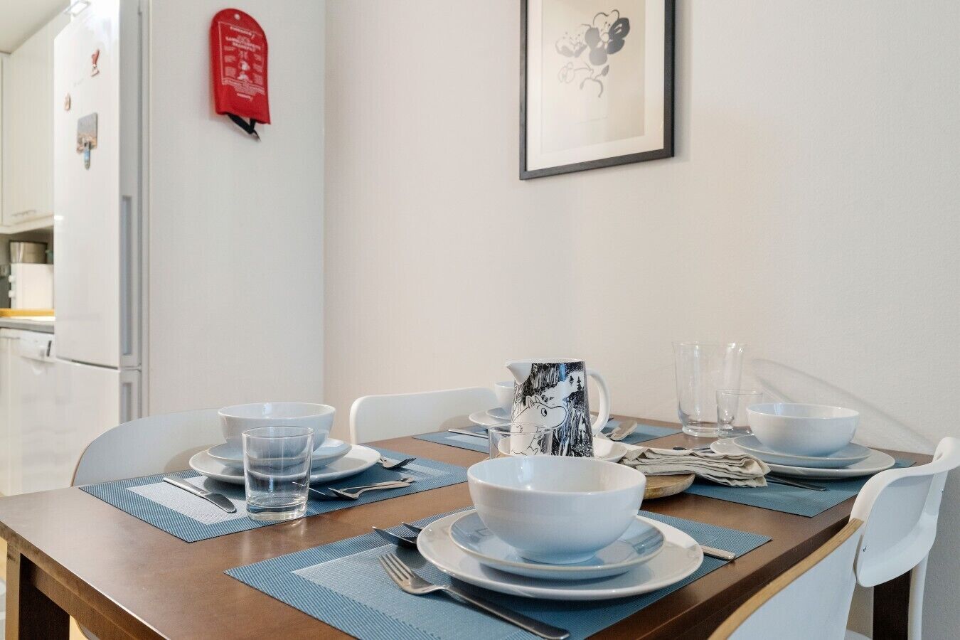 A dining table set with plates, bowls, glasses, and cutlery, with a framed picture on the wall and a fire extinguisher on the wall.