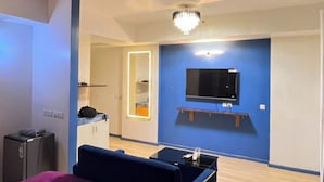 A bedroom with a bed, a ceiling fan, a TV mounted on the wall, and a small table. - Luxury Apartments Stay at DLF (Lucknow)