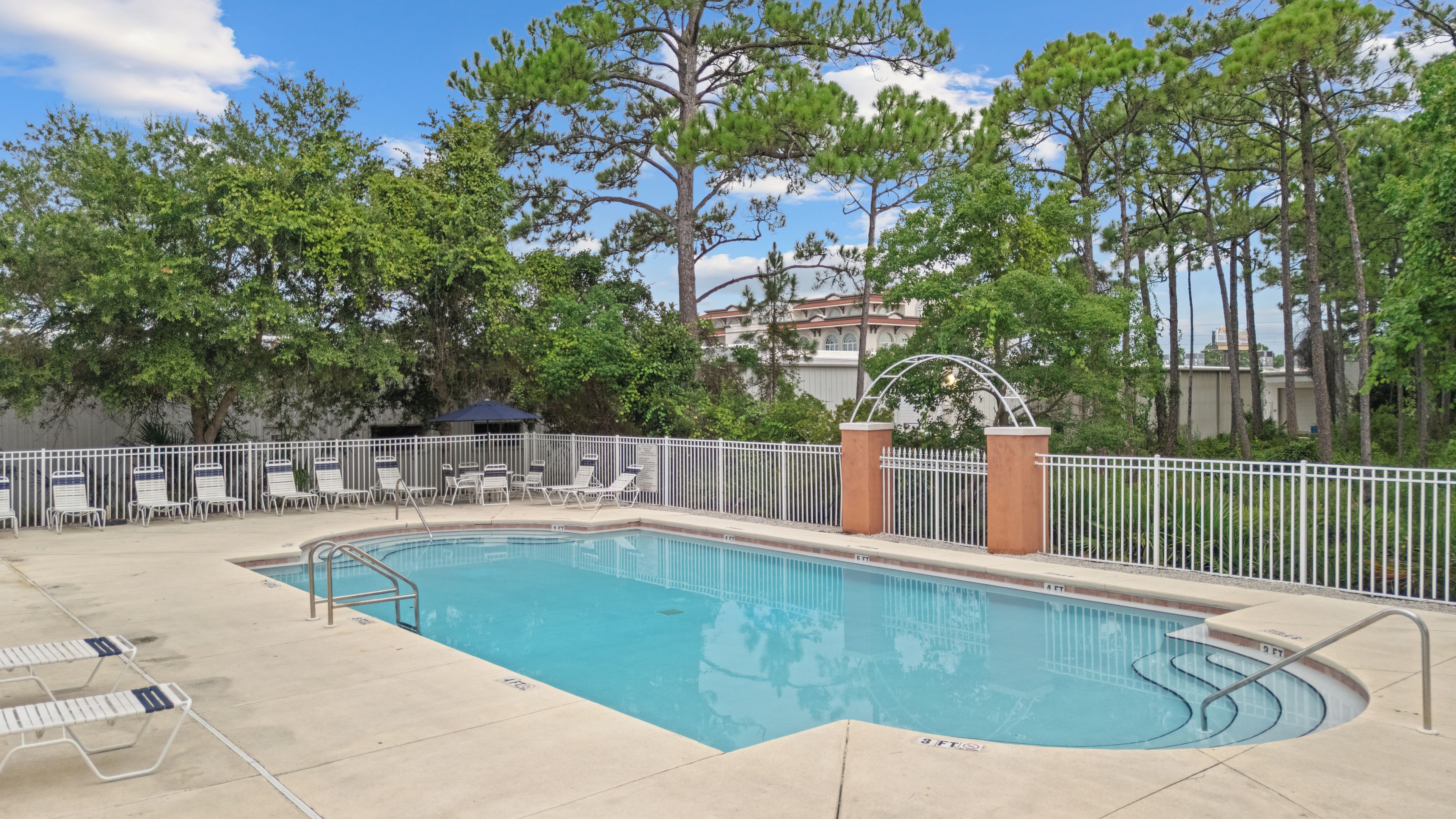 A rectangular swimming pool with a white metal fence, lounge chairs, and a covered seating area.