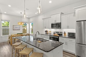 A modern kitchen with white cabinetry, a granite countertop, stainless steel appliances, and a central island with bar stools. - Golf Cart + Game Room + Pool Access | Steps to the Sand | Sundrift (Miramar Beach)