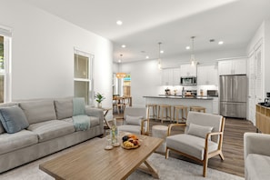 A modern living room with a sofa, chairs, a coffee table, and a kitchen in the background. - Golf Cart + Game Room + Pool Access | Steps to the Sand | Sundrift (Miramar Beach)