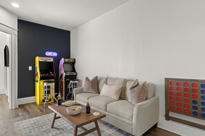 Ein Wohnzimmer mit einem beigefarbenen Sofa, einem Couchtisch, einer Pac-Man-Spielmaschine und einem GoSports-Spielbrett.