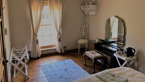 A bedroom with a piano, a chair, a bed, a window with curtains, a ceiling fan, and a shelf with decorative items.