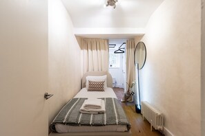 Ein schmales Hotelzimmer mit Bett, Heizkörper, Spiegel und einem Fenster mit Vorhängen.