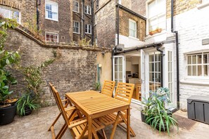 A wooden table and chairs set in a courtyard with brick walls and a white building in the background. - Stylish Bloomsbury Two-Bed Retreat (London)