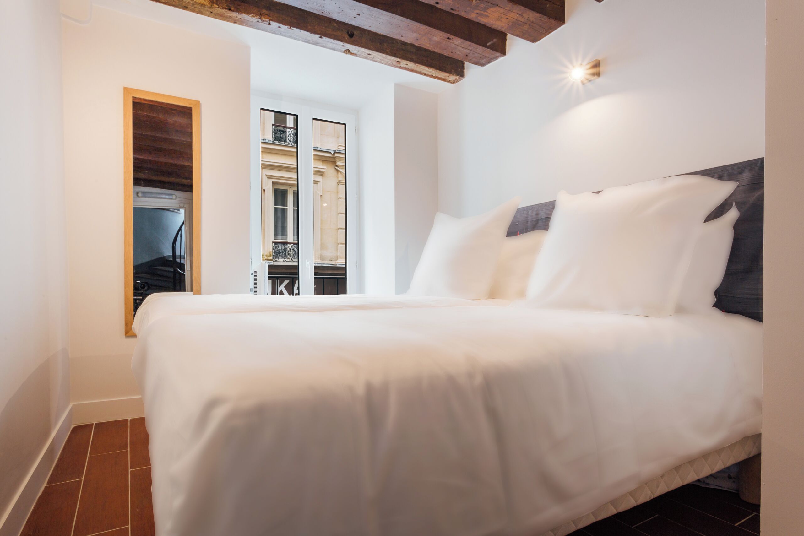 Une chambre avec un grand lit, une poutre en bois au plafond, une fenêtre donnant sur des immeubles et un luminaire fixé au mur.