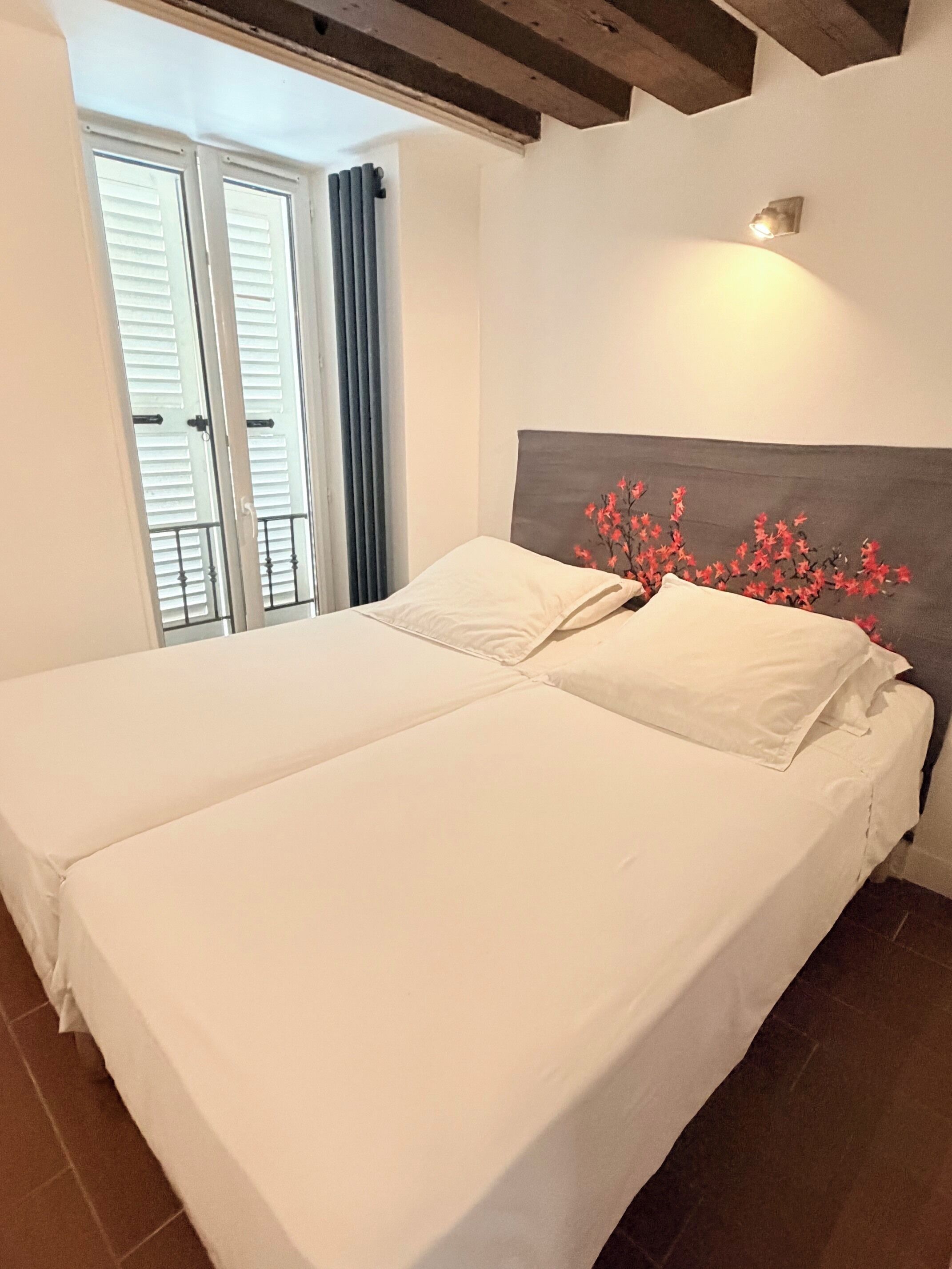 A bed with white linens and a decorative headboard, a window with blinds, and a wall-mounted light fixture.