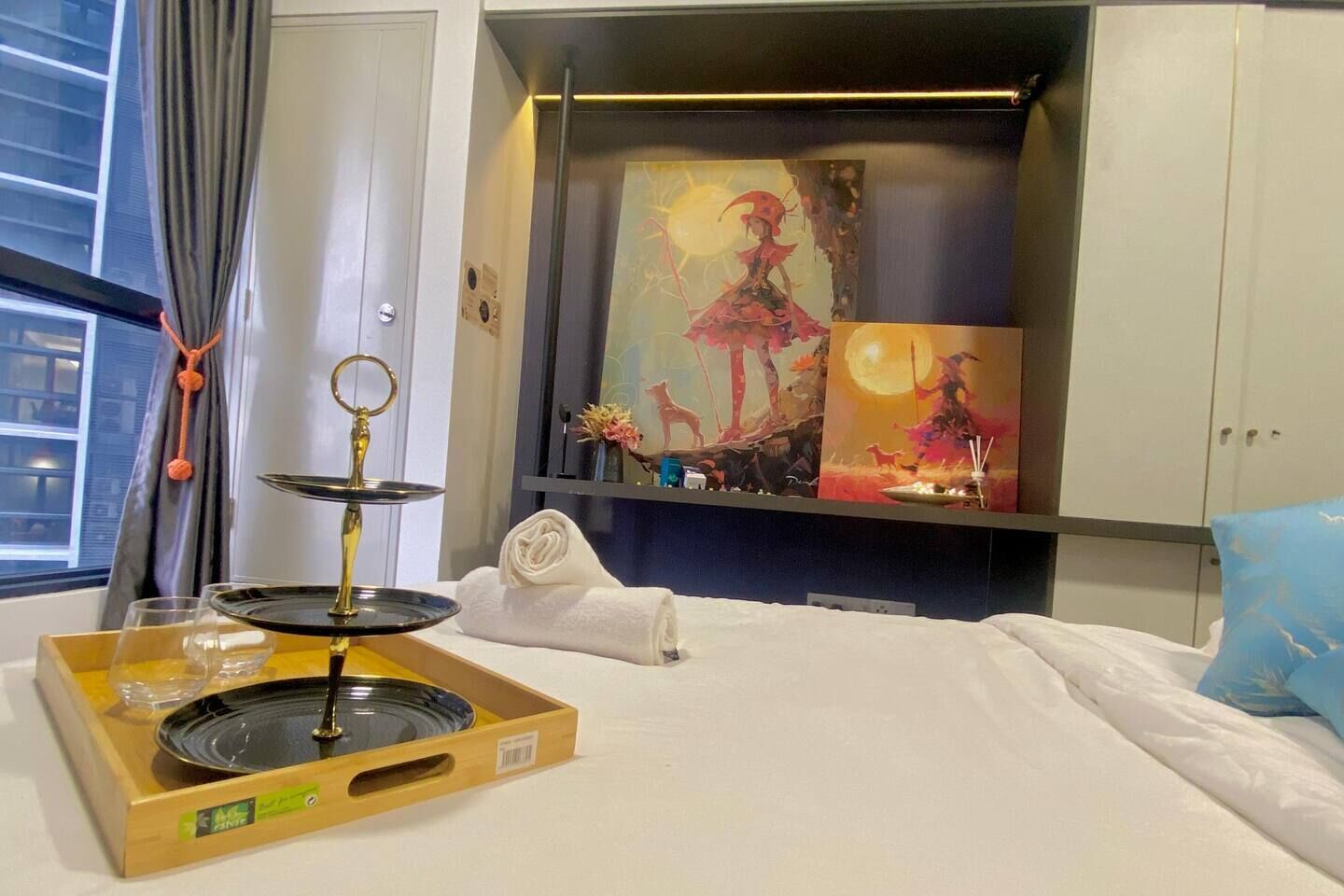 Ein Hotelzimmer mit einem Bett, einem Nachttisch mit Tablett, einem Bild an der Wand und einem Fenster mit Vorhängen.