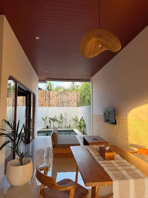 Área de refeições interna com vista para a piscina, móveis de madeira e luminária pendente.