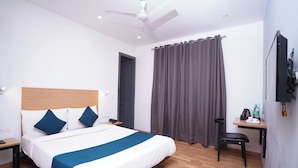 Quarto de hotel com cama, escrivaninha, cadeira, ventilador de teto e janela com cortinas.