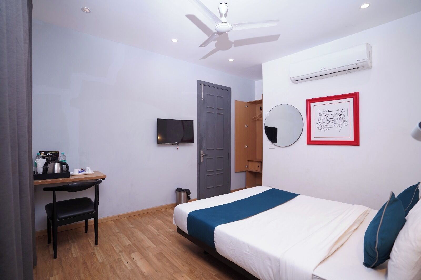 A hotel room with a bed, a desk with a chair, a television, a wardrobe, a mirror, and an air conditioning unit.
